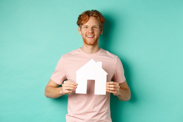 Foto gratuita concepto de bienes raíces. hombre joven con el pelo rojo, vestido con camiseta, mostrando el recorte de la casa de papel y sonriendo feliz, de pie sobre fondo de menta. copia espacio