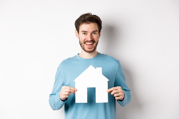 Concepto de bienes raíces y seguros. Hombre moderno guapo comprando una propiedad, sonriendo y mostrando el recorte de la casa de papel, de pie sobre fondo blanco.
