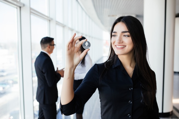 Concepto de blockchain e inversión. Líder de mujer de negocios sosteniendo litecoin frente a su equipo con las manos levantadas en la oficina.