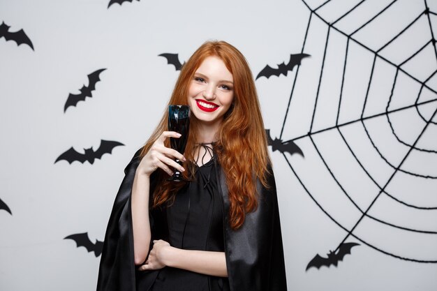 Concepto de bruja de Halloween - bruja feliz de Halloween sosteniendo un vaso de vino tinto con sangre sobre la pared gris oscuro con murciélago y tela de araña.