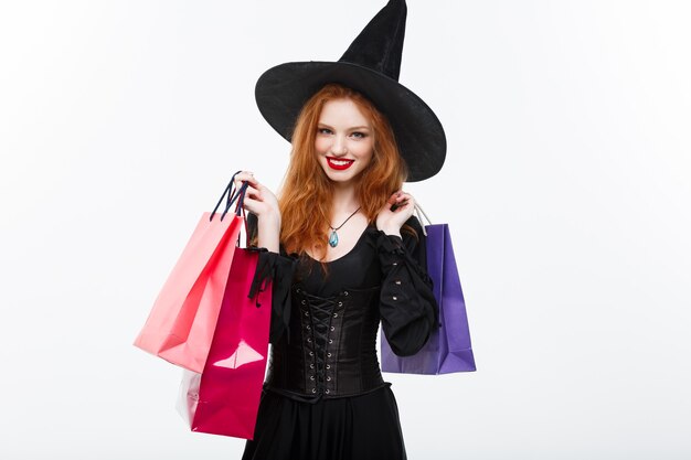 Concepto de bruja de Halloween bruja de halloween feliz sonriendo y sosteniendo coloridas bolsas de compras en la pared blanca