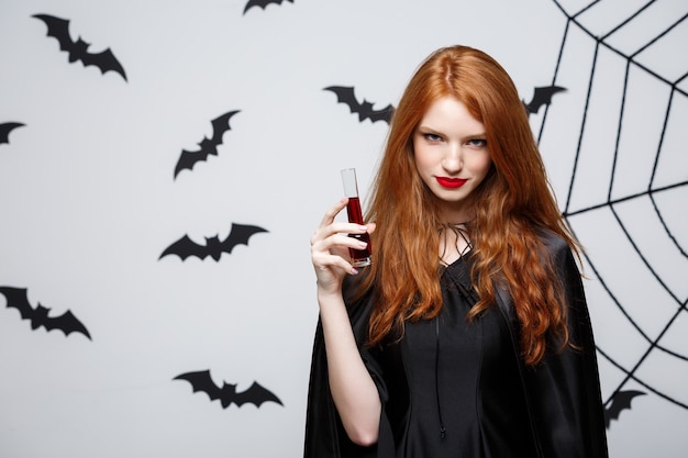 Concepto de bruja de Halloween - Feliz bruja de Halloween bebiendo sangre sobre una pared gris oscuro con murciélago y tela de araña.