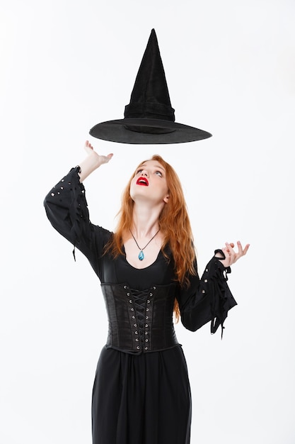 Concepto de bruja de Halloween - Feliz Halloween Bruja sexy de pelo de jengibre con sombrero mágico volando sobre su cabeza. Aislado en la pared blanca.