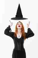 Foto gratuita concepto de bruja de halloween - feliz halloween bruja sexy de pelo de jengibre con sombrero mágico volando sobre su cabeza. aislado en la pared blanca.