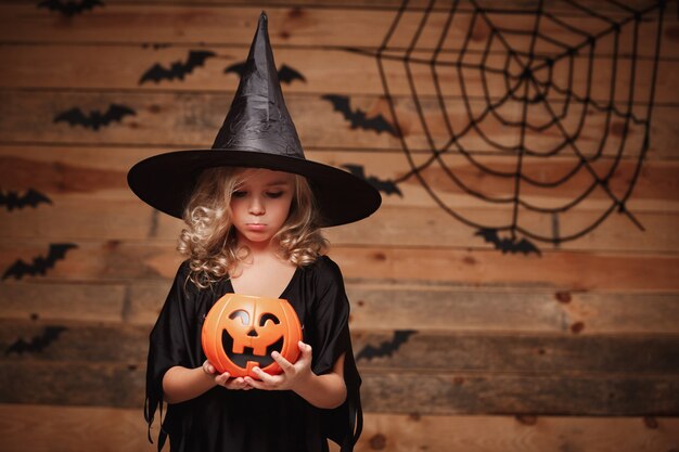 Concepto de bruja de Halloween - pequeño niño brujo caucásico decepcionante sin dulces en el tarro de calabaza de halloween. sobre fondo de telaraña y murciélago.