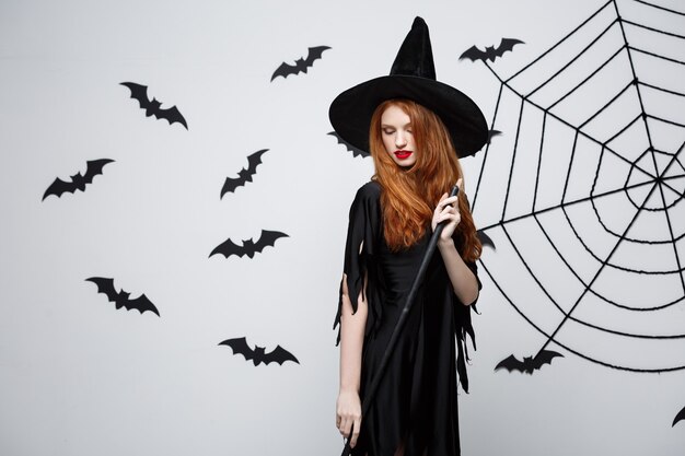 Concepto de bruja de Halloween retrato de joven y bella bruja con escoba sobre pared gris con pared de telaraña y murciélago