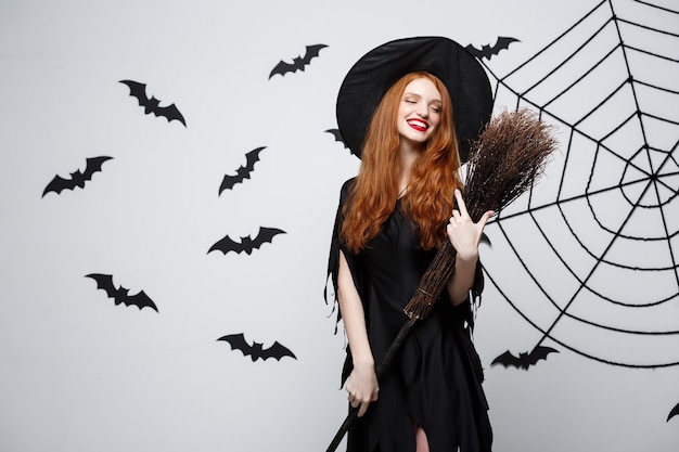Concepto de bruja de Halloween retrato de joven y bella bruja con escoba sobre pared gris con pared de telaraña y murciélago