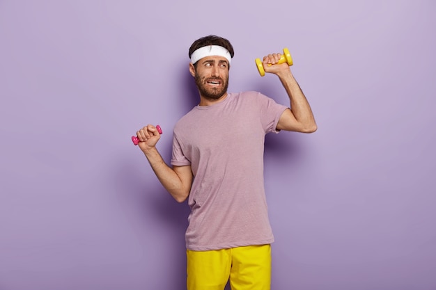 Concepto de cansancio y entrenamiento. Hombre sin afeitar disgustado levanta los brazos con mancuernas, siente fatiga de entrenamiento prolongado, vestido con ropa deportiva