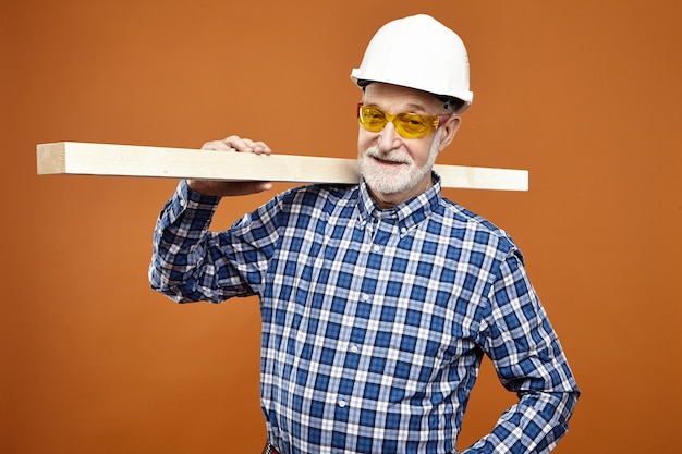 Foto gratuita concepto de carpintería, artesanía y carpintería. leñador o carpintero maduro senior positivo seguro con barba espesa sonriendo, llevando una tabla de madera sobre su hombro en la pared naranja en blanco