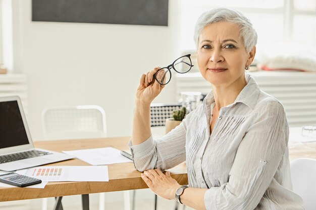 Concepto de carrera, negocios y éxito. Atractiva empresaria guapa en blusa gris sedosa sentada en su lugar de trabajo con computadora portátil, papeles y calculadora en el escritorio, sosteniendo gafas, teniendo descanso