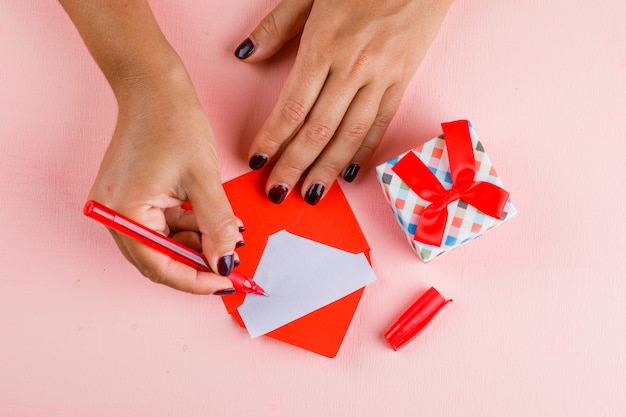 Concepto de celebración con caja de regalo en mesa rosa plana lay. mujer firma tarjeta de felicitación.