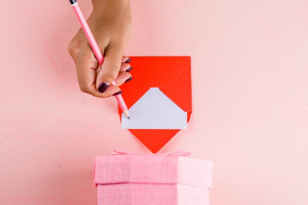 Concepto de celebración con caja de regalo en mesa rosa plana lay. mujer firma tarjeta de felicitación.