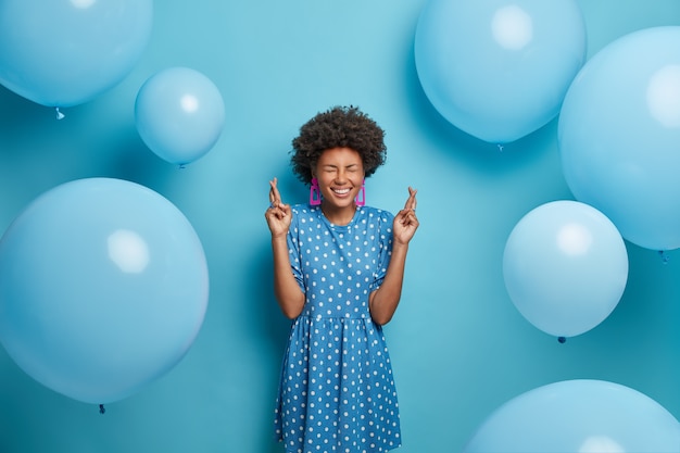 Concepto de celebración feliz. La cumpleañera positiva y esperanzada cruza los dedos, pide un deseo, cree que todos sus sueños se hacen realidad vestida con un vestido de lunares en un tono de pared. Globos inflados alrededor