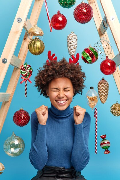 Concepto de celebración feliz. Positiva mujer de pelo rizado aprieta los puños se regocija de las buenas noticias vestida con un cuello de tortuga informal cuernos de reno posa contra el fondo azul con juguetes de año nuevo decora la habitación