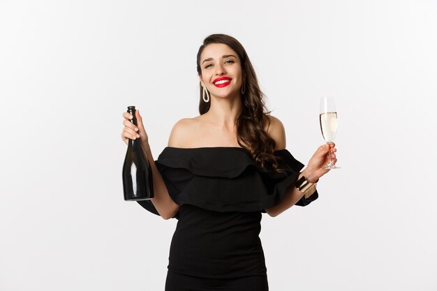 Concepto de celebración y fiesta. Elegante mujer morena vestida de glamour con botella y copa de champán, sonriendo complacido, de pie sobre fondo blanco.