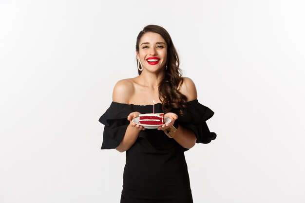 Concepto de celebración y fiesta. Feliz hermosa mujer teniendo cumpleaños, sosteniendo el b-day cake y sonriendo, pidiendo deseos, de pie en vestido negro con maquillaje.