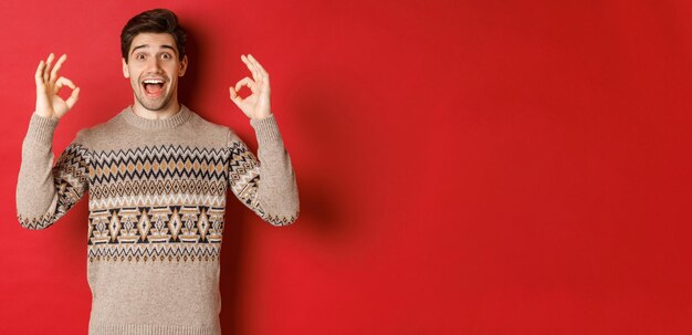 Concepto de celebración navideña, vacaciones de invierno y estilo de vida. Retrato de un hombre guapo con suéter, que se ve asombrado y muestra señales de estar bien, garantiza o recomienda algo bueno, fondo rojo.