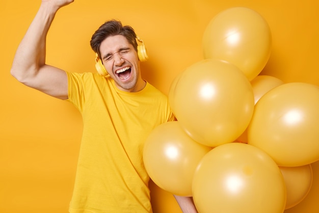 Concepto de celebración y personas El hombre positivo baila despreocupado exclama de alegría mantiene los ojos cerrados sostiene un montón de globos inflados escucha música a través de auriculares vestidos con una camiseta aislada en amarillo