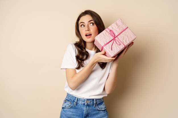 Concepto de celebración y vacaciones. Chica joven feliz que parece intrigada, sacudiendo la caja con el regalo, adivinando qué hay dentro del regalo envuelto, de pie sobre un fondo beige.