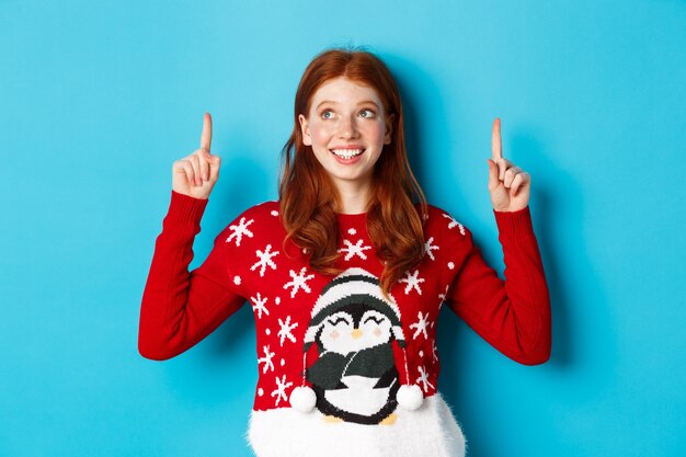 Concepto de celebración y vacaciones de invierno. Chica adolescente alegre con el pelo rojo, mirando soñadora en el logo, señalando con el dedo hacia arriba, mostrando publicidad, de pie sobre fondo azul.