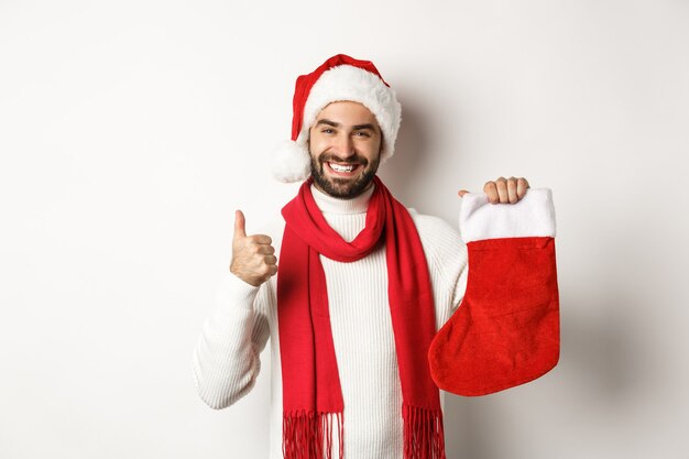 Concepto de celebración y vacaciones de invierno. Hombre feliz mostrando calcetín de Navidad para regalos y pulgar hacia arriba, sonriendo satisfecho, de pie sobre fondo blanco.
