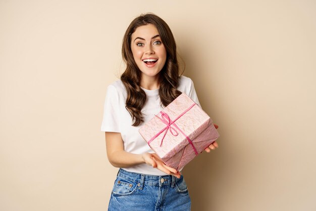 Concepto de celebración y vacaciones. Una joven feliz sosteniendo un regalo envuelto en una caja rosa, recibe un regalo, luciendo asombrada y sorprendida, de pie sobre un fondo beige.