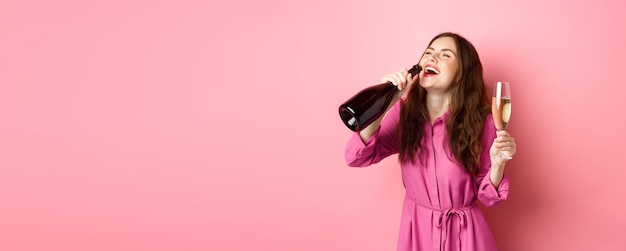 Foto gratuita concepto de celebración y vacaciones mujer despreocupada feliz bebiendo champán de una botella con una sonrisa complacida relajándose en la fiesta sosteniendo un vaso de pie sobre fondo rosa