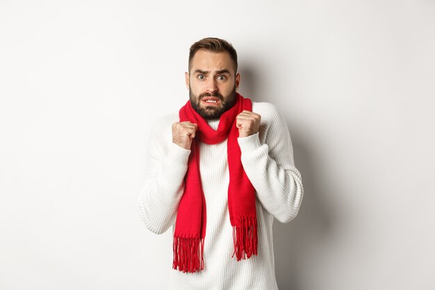 Concepto de celebración y vacaciones de Navidad. Hombre asustado temblando de miedo y mirando sorprendido, de pie ansioso con bufanda roja y suéter blanco