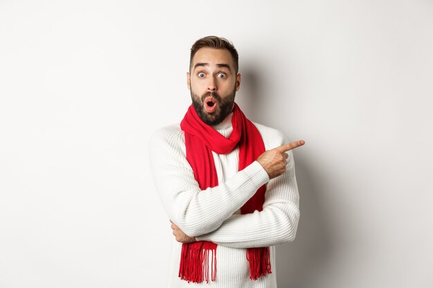 Concepto de celebración y vacaciones de Navidad. Hombre barbudo sorprendido apuntando con el dedo hacia la derecha y mirando impresionado a la cámara, de pie con bufanda roja y suéter, fondo blanco.