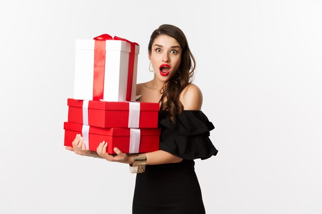 Concepto de celebración y vacaciones navideñas. Hermosa mujer en vestido negro sosteniendo regalos y mirando sorprendido, de pie sobre fondo blanco.