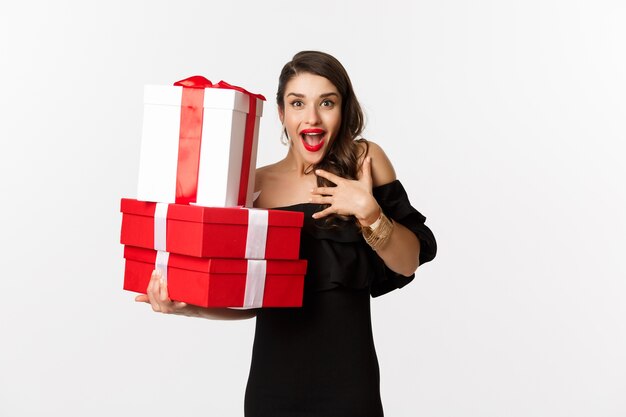 Concepto de celebración y vacaciones navideñas. Mujer emocionada y feliz recibe regalos, sosteniendo regalos de Navidad y regocijándose, de pie en vestido negro sobre fondo blanco.