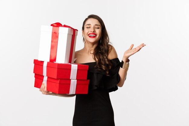 Concepto de celebración y vacaciones navideñas. Mujer emocionada y feliz recibe regalos, sosteniendo regalos de Navidad y regocijándose, de pie en vestido negro sobre fondo blanco.