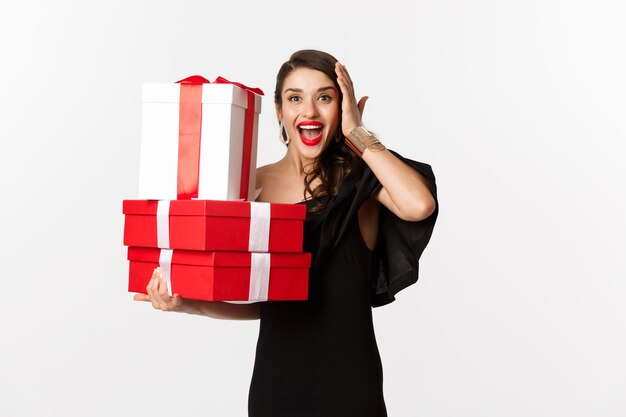 Concepto de celebración y vacaciones navideñas. Mujer emocionada y feliz recibe regalos, sosteniendo regalos de Navidad y regocijándose, de pie en vestido negro sobre fondo blanco.