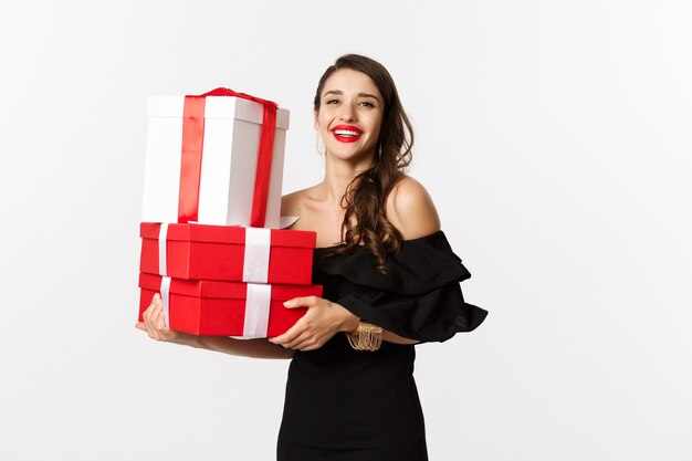 Concepto de celebración y vacaciones navideñas. Mujer de moda en vestido negro elegante, sosteniendo regalos y sonriendo, de pie sobre fondo blanco.