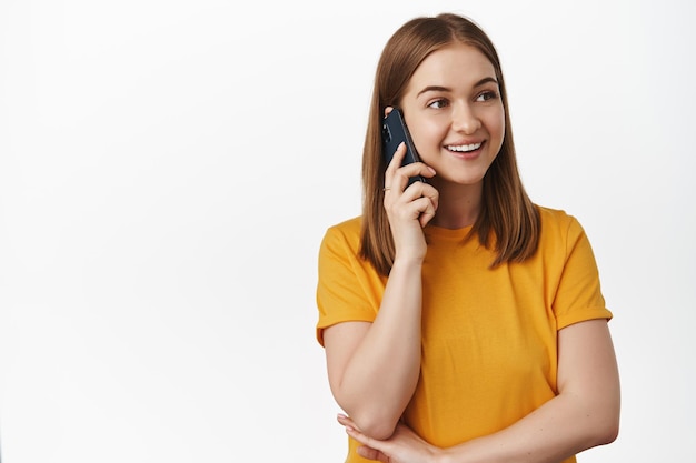 Concepto celular y tecnológico. Mujer joven hablando por teléfono móvil y sonriendo, vistiendo camiseta amarilla. Chica hablando con un amigo y mirando a un lado, usando un teléfono inteligente, fondo blanco