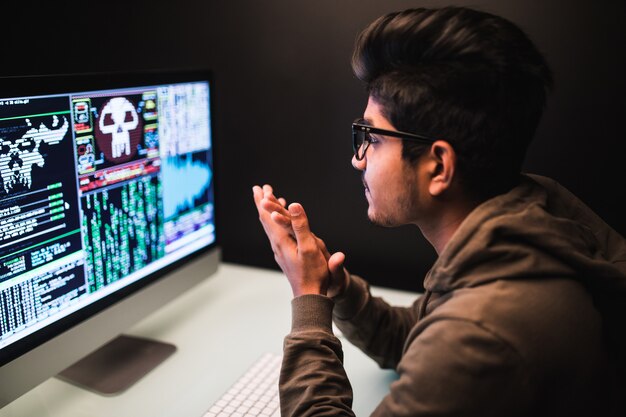Concepto de ciberdelincuencia, piratería y tecnología: hacker masculino joven con teléfono inteligente y codificación en la pantalla de la computadora en un cuarto oscuro