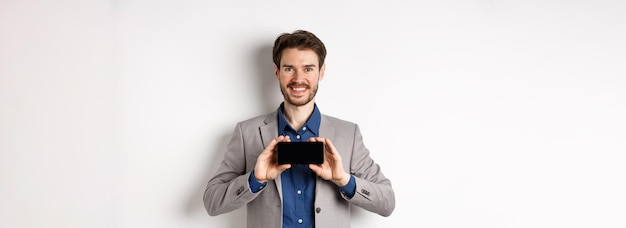 Foto gratuita concepto de comercio electrónico y compras en línea chico feliz en traje de negocios que muestra la pantalla del teléfono inteligente vacía hor