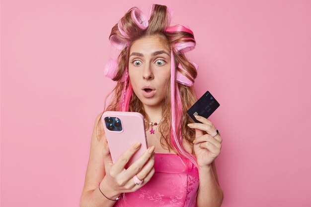 Concepto de comercio electrónico y compras en línea Una joven impresionada mira fijamente el teléfono inteligente sostiene una tarjeta de crédito de plástico y aplica rodillos para el cabello para hacer que el peinado use un vestido aislado sobre un fondo rosa