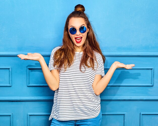 Concepto de comparación. Joven mujer morena en ropa casual de verano hipster mostrando algo en ambas manos planas para una elección similar de producto, posando cerca de la pared azul