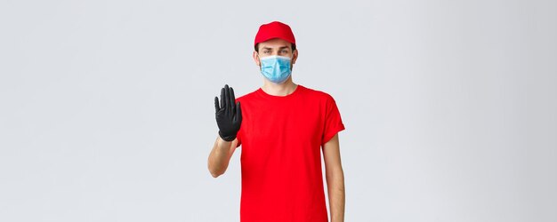 Concepto de compra en línea de autocuarentena de covid19 Un repartidor confiado con guantes de uniforme rojo y mascarilla levanta la mano en la señal de alto para evitar que el cliente pase al almacenamiento sin equipo de protección