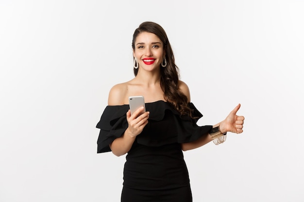 Concepto de compra online. Mujer atractiva en vestido negro de moda, maquillaje, mostrando el pulgar hacia arriba y usando la aplicación de teléfono móvil, fondo blanco.
