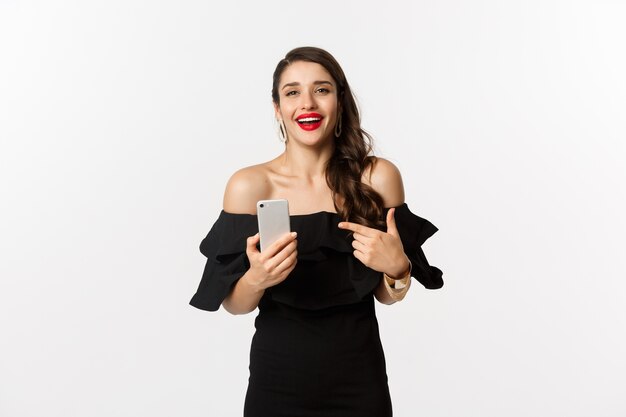 Concepto de compra online. Mujer bonita satisfecha en vestido negro, sonriendo complacido y apuntando al teléfono móvil, de pie sobre fondo blanco.