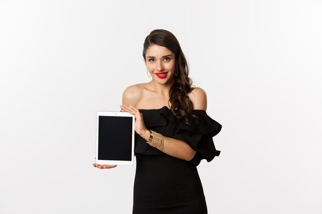 Concepto de compra online. Mujer bonita tentada en vestido negro que muestra la pantalla de la tableta digital, de pie sobre fondo blanco. Copia espacio