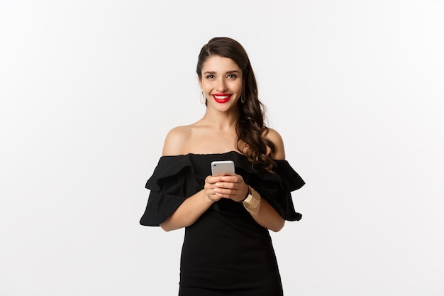 Concepto de compra online. mujer joven vestida de negro, leyendo un mensaje de texto, usando el teléfono móvil y sonriendo, de pie sobre fondo blanco.