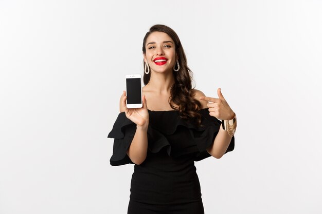 Concepto de compra online. Mujer de moda en vestido negro apuntando con el dedo a la pantalla del teléfono inteligente, mostrando la aplicación, de pie sobre fondo blanco.