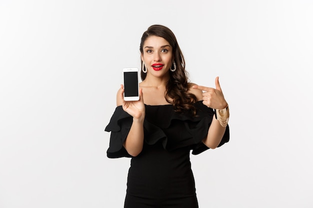 Concepto de compra online. Mujer de moda en vestido negro apuntando con el dedo a la pantalla del teléfono inteligente, mostrando la aplicación, de pie sobre fondo blanco.