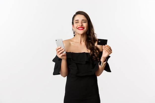 Concepto de compra online. Mujer de moda en vestido negro, sosteniendo la tarjeta de crédito con el teléfono inteligente, mirando satisfecho, de pie sobre fondo blanco.