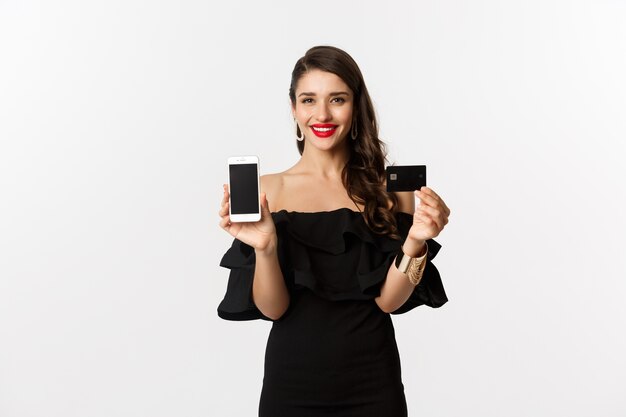 Concepto de compra online. Mujer morena de moda en vestido negro, mostrando la pantalla del móvil y la tarjeta de crédito, sonriendo complacido, de pie sobre fondo blanco.