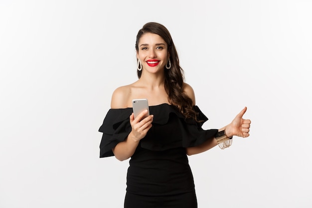 Concepto de compra online. mujer en vestido negro de moda, maquillaje, mostrando el pulgar hacia arriba y usando la aplicación de teléfono móvil, fondo blanco.