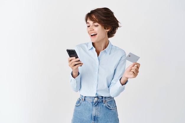 Concepto de compras y comercio electrónico Feliz joven europea haciendo un pedido en línea con tarjeta de crédito y teléfono móvil mirando la pantalla y fondo blanco sonriente